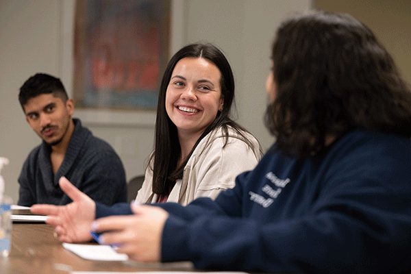 Three students sit at a desk in a classroom. On the right is a male student with black hair, wearing a grey sweater. He is looking at the other students. In the center is a female student with long brown hair. She wears a tan jacket and is smiling at the student on the left with short brown hair, wearing a blue sweatshirt. A painting is on the wall behind them.