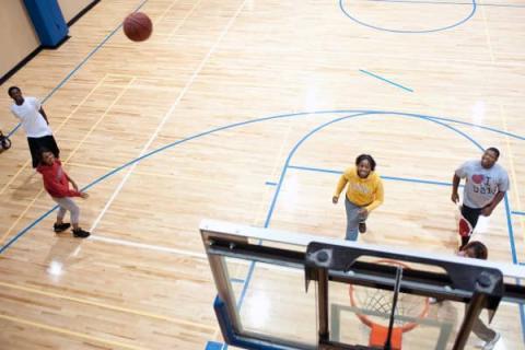 A group of five Black individuals playing basketball on a basketball court. The photo is taken from above the basketball hoop. The players wear a variety of white, red, yellow, grey, and black informal athletic gear (shorts, sweats, T-shirts, etc.). The basketball is visible mid-air, going towards the basketball hoop. The other players are looking up at the hoop.