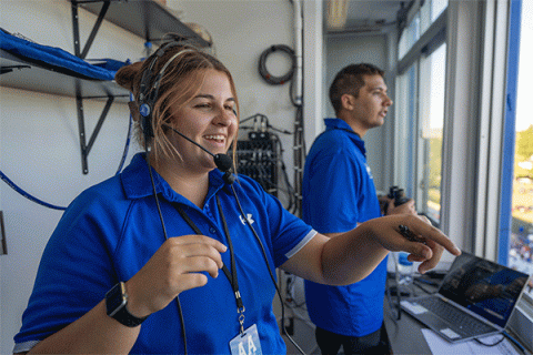 A female student wearing a blue shirt pointing out of the window of a press box with a male student in blue shirt in the background.